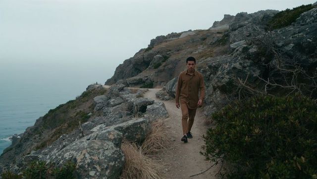 Walking asian man on coastal cliff trail wearing brown outfit overlooking misty ocean