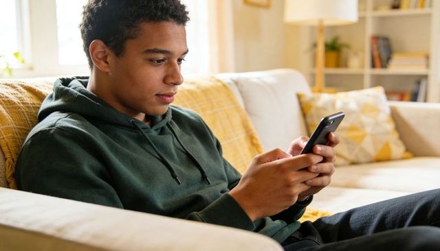 Teen sitting on sofa using smartphone in relaxed cozy living room with dark green hoodie