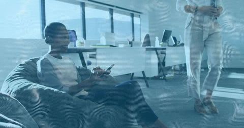 Professional scrolling phone on beanbag while coworker holds tablet in bright workspace