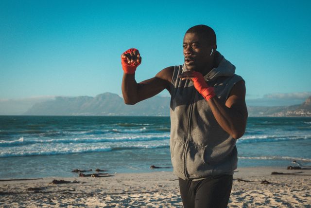 African american man exercising, shadowboxing with wrapped hands on beach at sunset. healthy outdoor lifestyle fitness training.