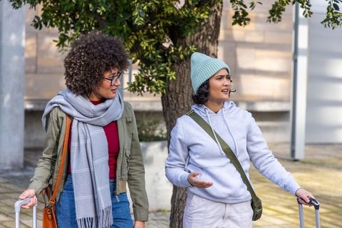 Two women walking and chatting with rolling luggage on urban sidewalk wearing hat and scarf
