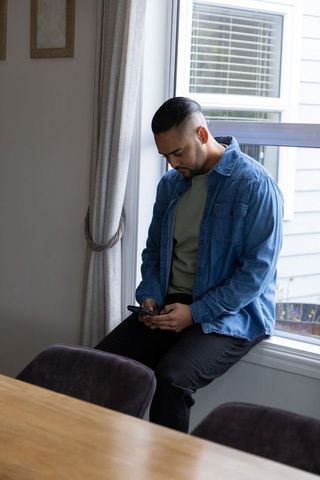 Young man using smartphone sitting on window ledge in modern dining nook, casual denim