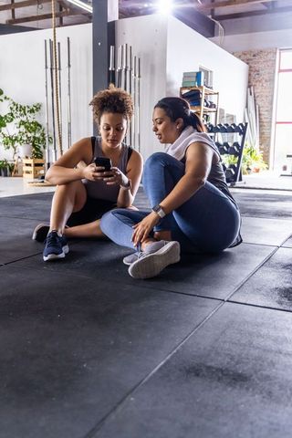 Female friends sharing smartphone and towel after workout in bright gym studio