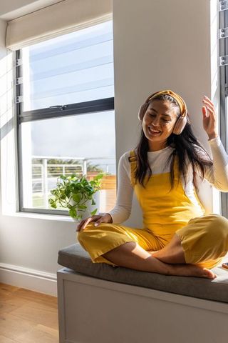 Sunlit asian woman relaxing on window seat wearing yellow overalls and pink headphones