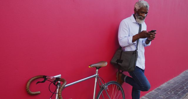 Bright and lively urban scene of a smiling senior man leaning against a vibrant red wall while checking his smartphone, with a bicycle by his side. Ideal for use in lifestyle blogs, technology ads targeting seniors, and urban living promotions.