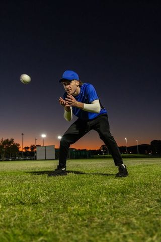 Catching baseball at dusk: male athlete crouching on grass under stadium floodlights