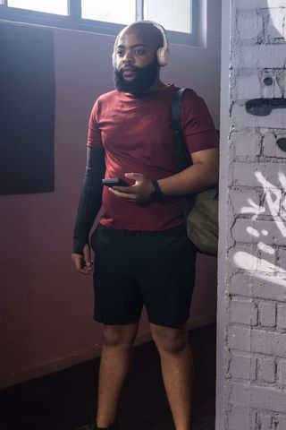 African man standing by graffiti column in corridor wearing headphones holding smartphone