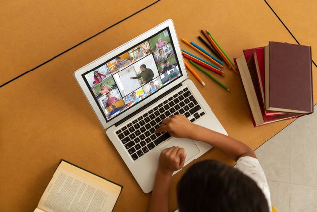 This image depicts a child engaged in online learning via a video call on a laptop. Books, colored pencils, and other school supplies are within reach. This visual is ideal for illustrating remote education, online schooling, and digital learning environments. Suitable for educational blogs, e-learning platforms, virtual classroom promotions, and articles about technology integration in education.