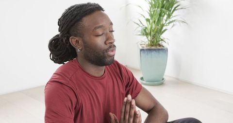 African American man meditating with prayer hands beside potted plant in minimal home