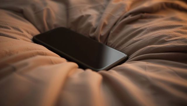 Black smartphone resting in beige quilted duvet crease reflecting warm morning light