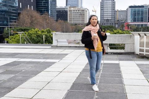 Asian woman walking with coffee and smartphone on urban terrace with skyline background