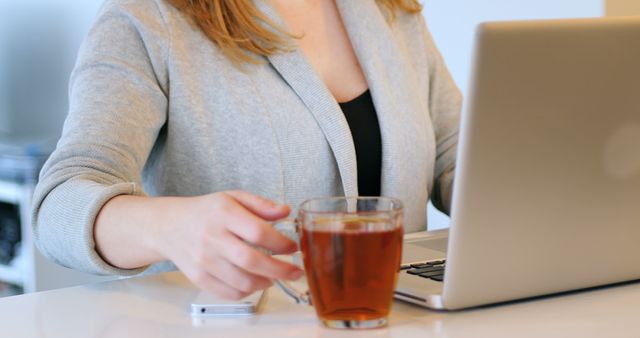 Woman focused on her laptop while drinking tea in a modern home office. Useful for illustrating remote work, home office setups, business lifestyle and balancing work with relaxation.