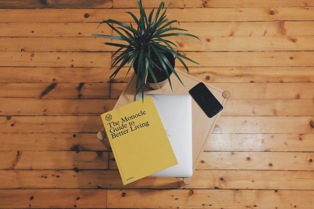 A table displaying a book titled 'The Monocle Guide to Better Living', a closed laptop, a potted plant, and a smartphone on a wooden floor. Ideal for use as a representation of a minimalist home office or workspace setup. Suitable for lifestyle, productivity, and interior decor content.