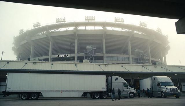 Foggy stadium looming over loading docks with tractor-trailers and crew preparing gear