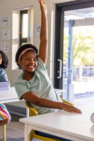 African american girl smiling raising hand in bright classroom by glass door with yellow chair