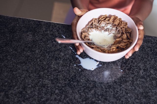 Hands of african american girl holding bowl with healthy cereals and milk on table for breakfast. Unaltered, food, morning, childhood, lifestyle and home concept.