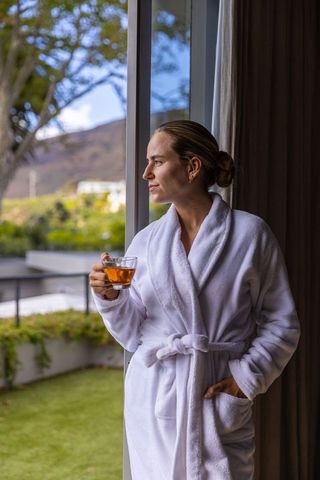 Woman relaxing in white bathrobe sipping tea by glass door with garden view