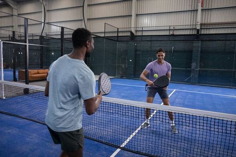 Diverse male players playing padel on indoor blue court hitting neon ball over net
