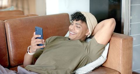African american man in 20s reclining on leather sofa, smiling while using smartphone