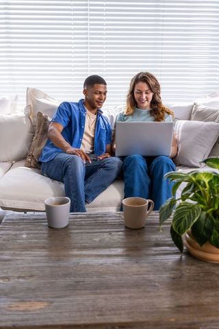 Diverse young couple working together on laptop on sofa in bright cozy living room