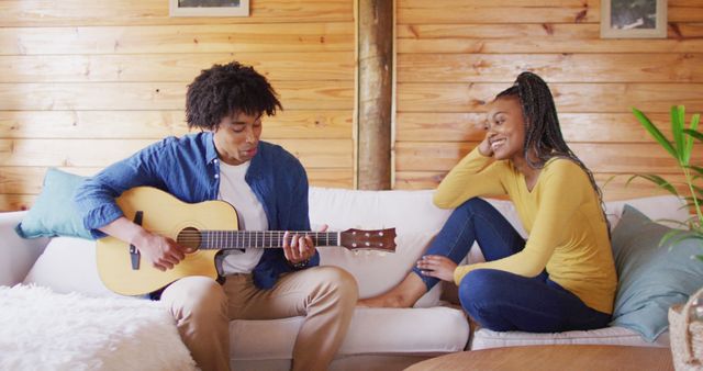 Happy african american couple playing guitar, sitting on sofa in log cabin, slow motion. Lifestyle, domestic life, countryside and nature concept.