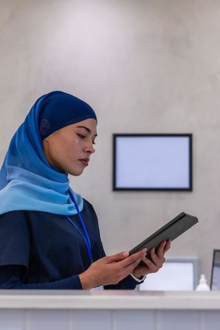 African american healthcare receptionist wearing blue hijab and scrubs using tablet at front desk