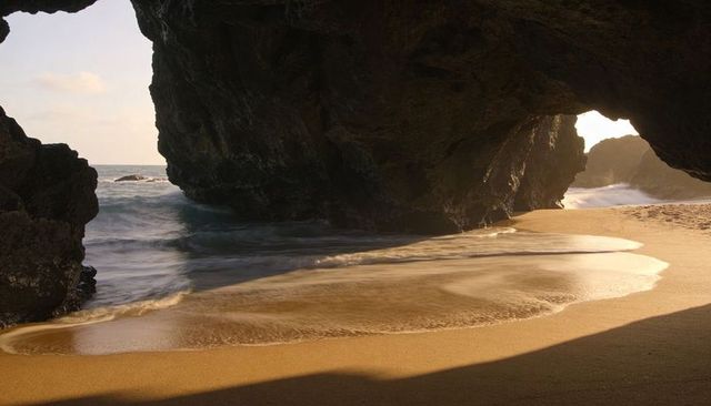 Sunlit sea cave framing golden wet sand and smooth tide under backlit coastal arch