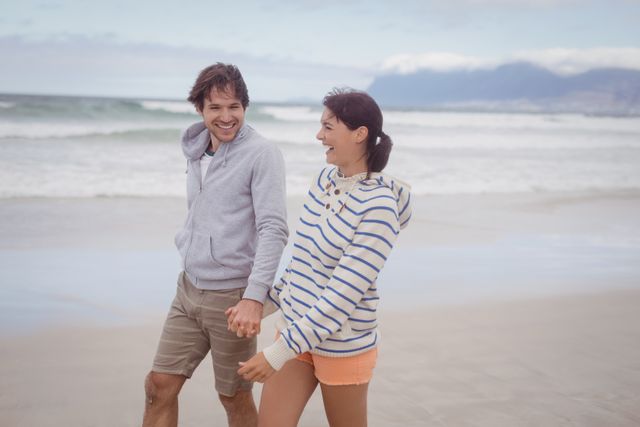 This image shows a happy young couple holding hands and walking along a beach. They are dressed in casual clothing, enjoying a leisurely stroll by the ocean. This image can be used for promoting travel destinations, romantic getaways, lifestyle blogs, or advertisements focused on outdoor activities and vacations.