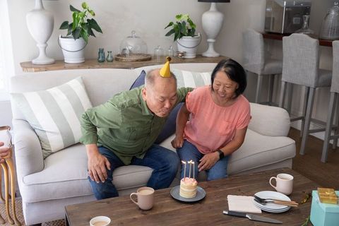 Asian senior couple celebrating intimate birthday at home man blowing candles on cake