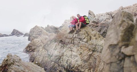 Mature couple hiking on rocky coastal cliff wearing red jackets and neon green backpack