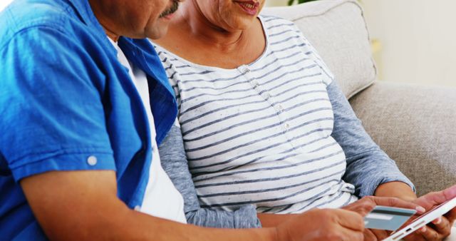 Senior couple sitting together at home, using a tablet. Both are focused on the screen, engaging with the technology. Ideal for subjects like elder technology use, digital inclusion, bonding, and modern lifestyle.