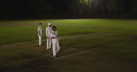 Night cricket teammates standing at batting crease in white kit with helmet and bat