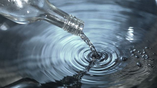 Clear water flowing from glass bottle creates splashing ripples in calm pool. Ideal for use in hydration-related advertisements, product campaigns for bottled water or purified liquids, and environmental videos or articles highlighting clean water.