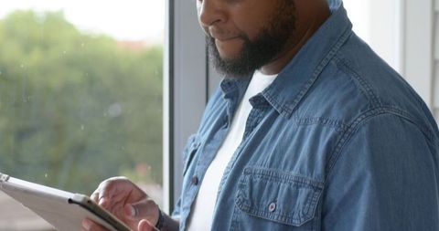 Man tapping tablet by window in denim shirt, thoughtful business lifestyle portrait