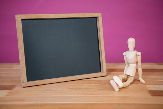 Wooden figurine sitting near a blank chalkboard on a wooden floor with a pink background. Ideal for educational themes, teaching concepts, creative projects, or messages. Can be used for presentations, blog posts, or social media content related to learning, education, or creativity.