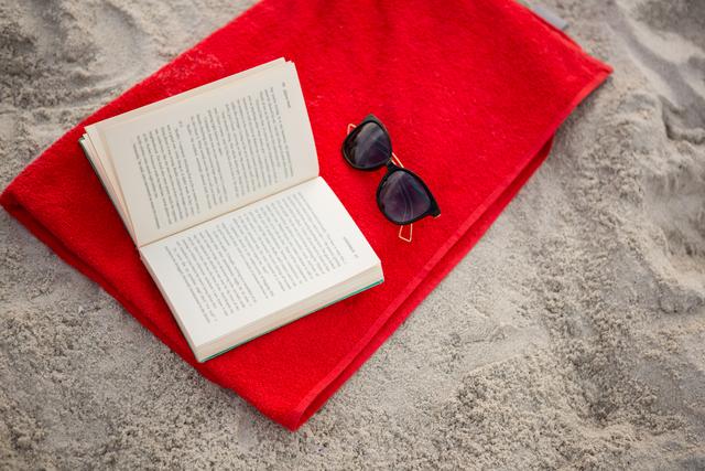 Open book and sunglasses placed on a red towel on sandy beach. Ideal for illustrating concepts of relaxation, summer vacations, leisure reading, and beach holidays. Suitable for travel blogs, vacation advertisements, and lifestyle articles.