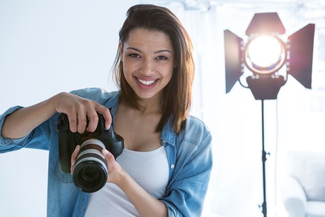 Young female photographer smiling while holding camera in a well-lit studio. Ideal for use in articles or advertisements related to photography, creative professions, studio setups, and professional portraits. Can also be used for promoting photography courses, workshops, or equipment.