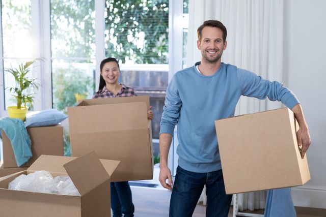 Young couple smiling while carrying cardboard boxes in their new home. Ideal for use in articles or advertisements about moving, relocation services, new homeownership, or lifestyle blogs focusing on young adults starting a new chapter in their lives.