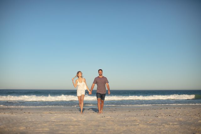Couple walking hand in hand on beach with ocean waves in background. Ideal for use in travel brochures, romantic getaway promotions, lifestyle blogs, and advertisements focusing on summer vacations, love, and relaxation.