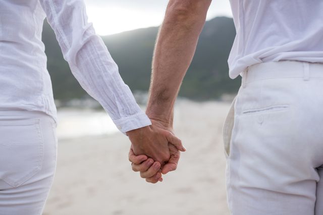 Couple holding hands while standing on a beach, dressed in white clothing. Ideal for themes of love, romance, relationships, summer vacations, and peaceful moments. Suitable for use in advertisements, travel brochures, wedding invitations, and lifestyle blogs.