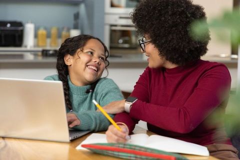 Caregiver helping girl with homework at kitchen table using laptop and notebook, smiling