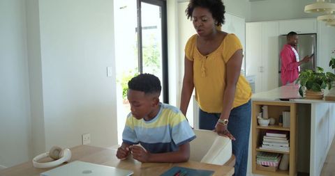 African american mother supervising son doing homework on laptop in open-plan kitchen