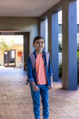 Asian schoolgirl standing in sunlit campus corridor wearing denim jacket and pink backpack
