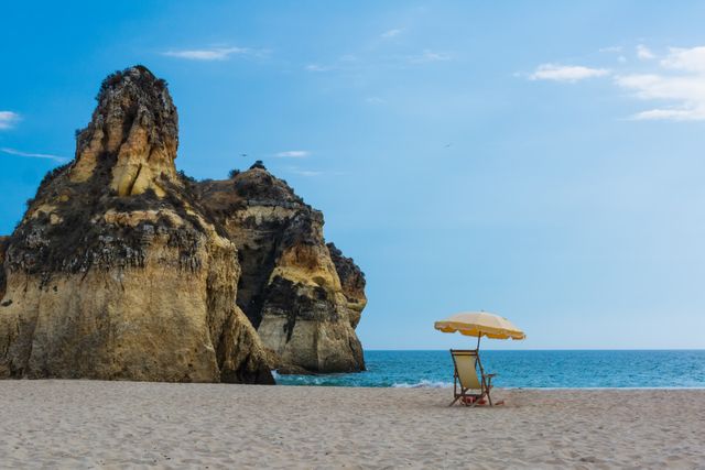 This serene beach scene captures an empty chair with a yellow umbrella near impressive rocky cliffs by the ocean. Great for promoting travel destinations, beach vacations, relaxation, and tranquil settings.