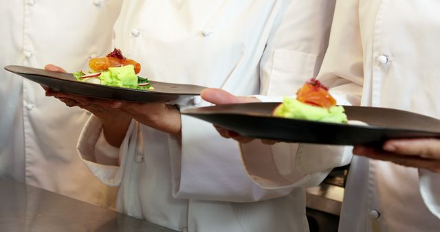 Three chefs in white uniforms are showcasing gourmet dishes in a professional kitchen. Each holds a stylish black plate with delicately plated food, emphasizing attention to detail and sophistication. This image is ideal for articles or advertisements regarding high-end dining, culinary schools, chef training programs, restaurant promotions, and food presentation guides.