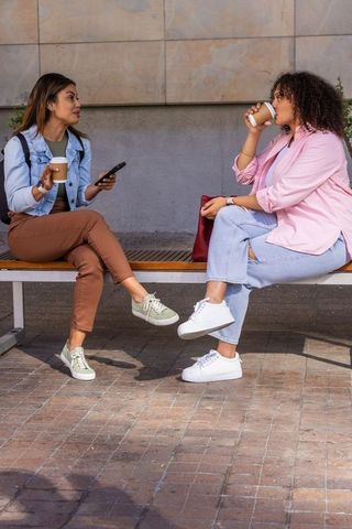 African american friends chatting and sipping coffee on bench in sunny urban plaza with phone