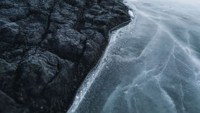 Dark jagged rock meeting frozen lake with thin ice rim and cracked veins