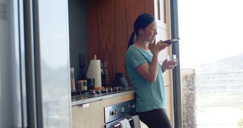 Asian woman leaning in modern kitchen speaking into smartphone holding glass on terrace