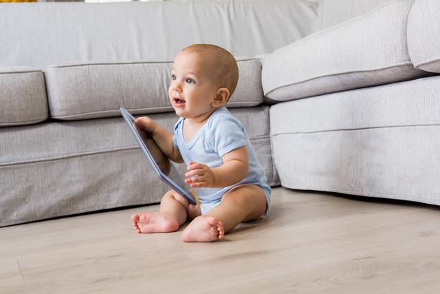 Baby boy sitting on floor in living room, holding digital tablet. Ideal for use in articles about childhood development, technology use among children, parenting tips, or home life. Suitable for blogs, educational materials, and advertisements related to baby products or tech gadgets.