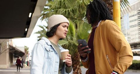 Friends discussing smartphone while holding coffee on urban sidewalk wearing mustard jacket